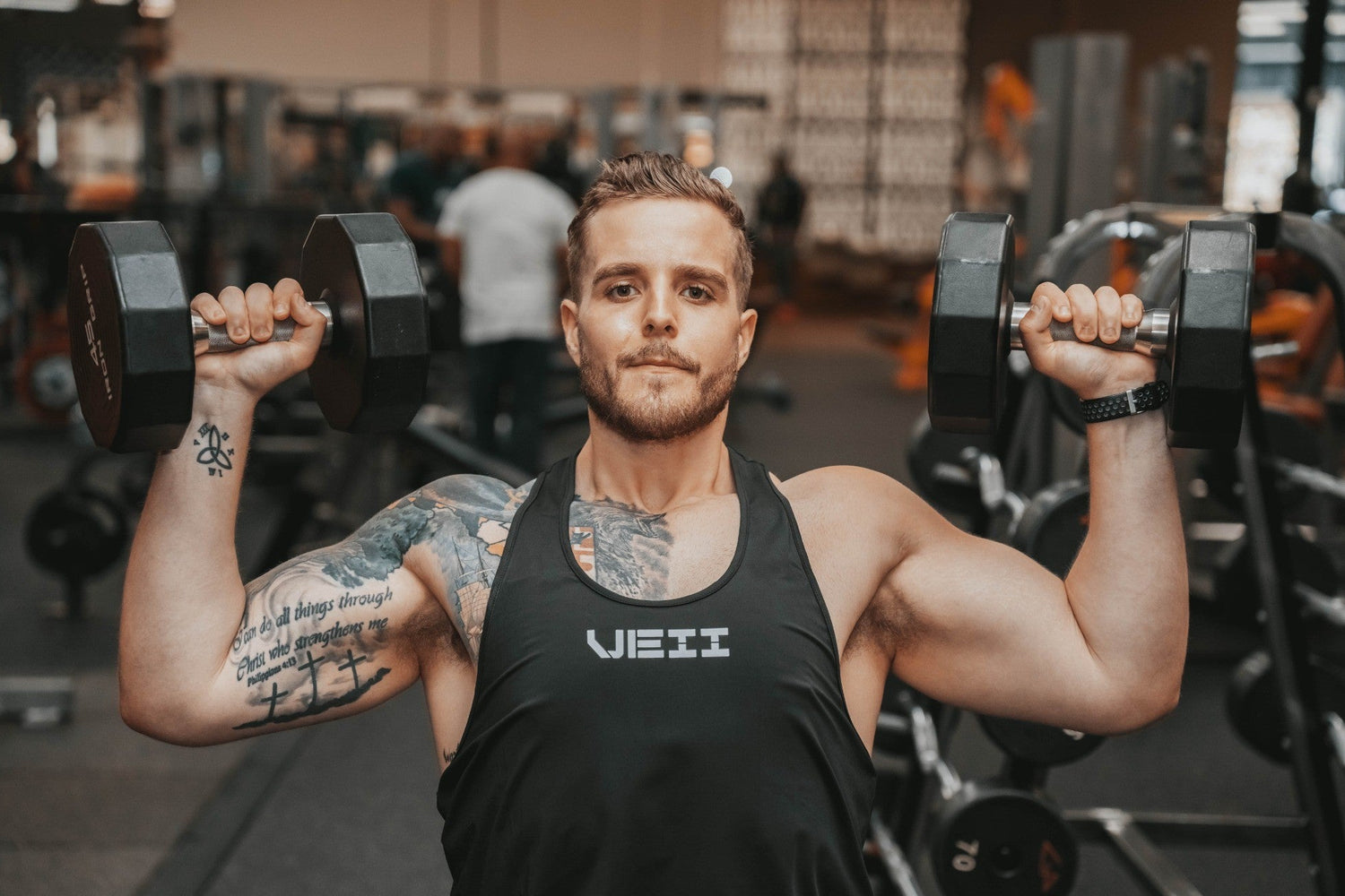 Athletic male trainer performing a dumbbell shoulder press in a gym, showcasing strength and control. Personalized 1:1 coaching available for customized workouts and expert guidance.